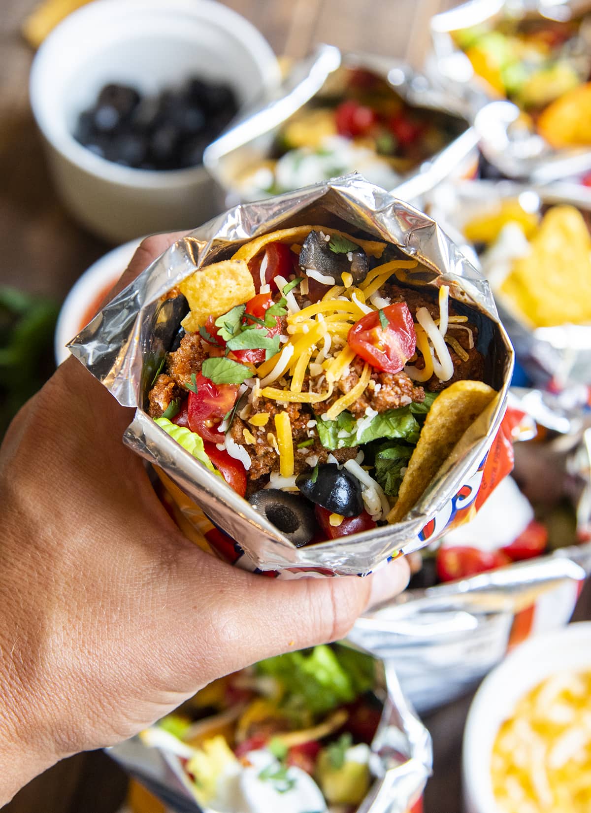 A walking taco held in a hand, you can see it is topped with chips, olives, tomatoes, cheese, and cilantro.