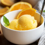 A bowl of scoops of orange sorbet with an orange slice and a mint leaf.