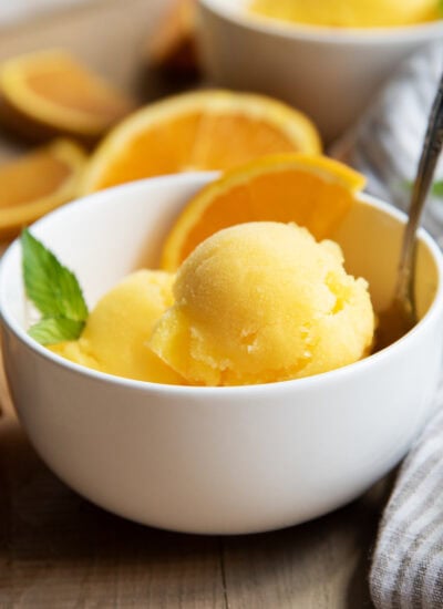 A bowl of scoops of orange sorbet with an orange slice and a mint leaf.