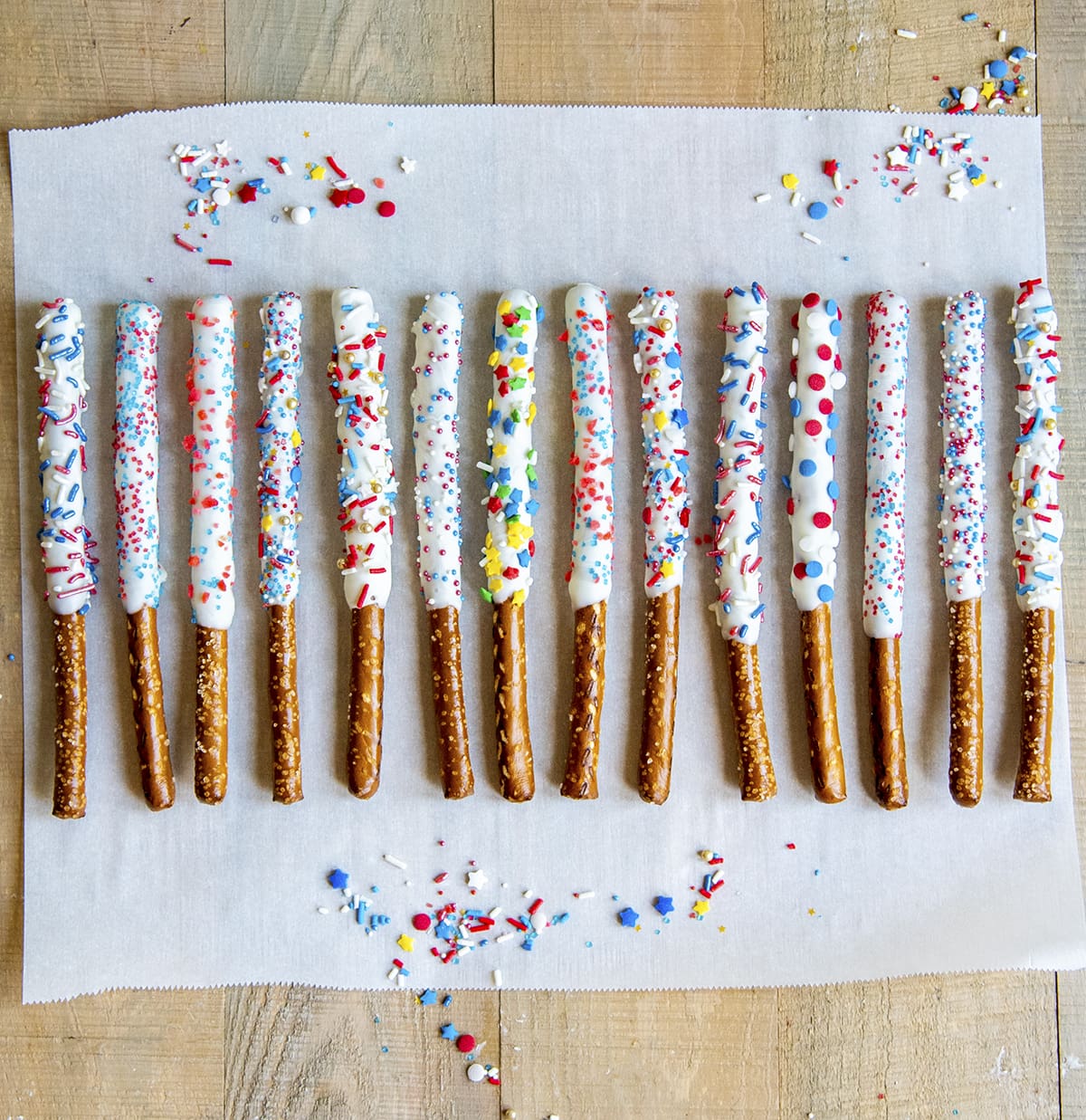 Pretzel rods dipped in white chocolate and topped with patriotic sprinkles on a piece of parchment paper.