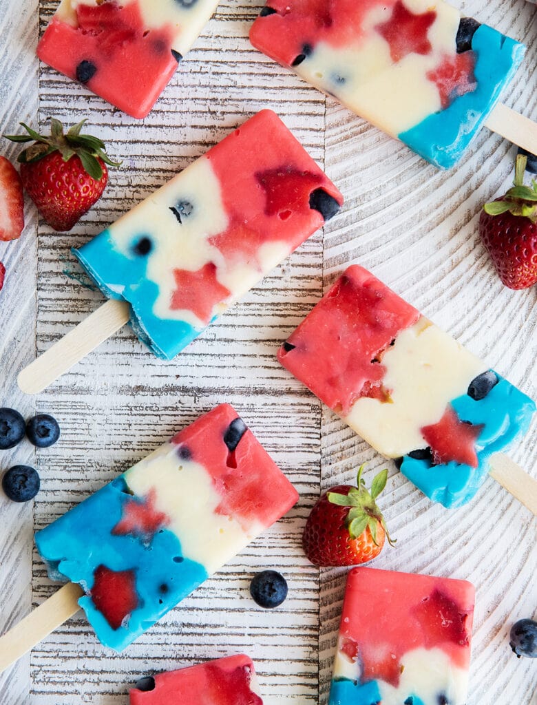 An above photo of red white and blue colored popsicles full of blueberries and star shaped strawberries on a white table.