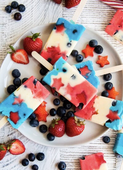 A plate of pudding popsicles that are colored red, white, and blue on a plate with blueberries and strawberries.