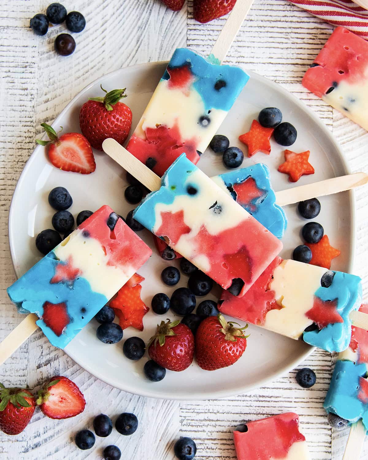A plate of pudding popsicles that are colored red, white, and blue on a plate with blueberries and strawberries.