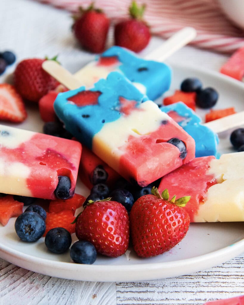 A plate of pudding popsicles that are colored red, white, and blue on a plate with blueberries and strawberries.