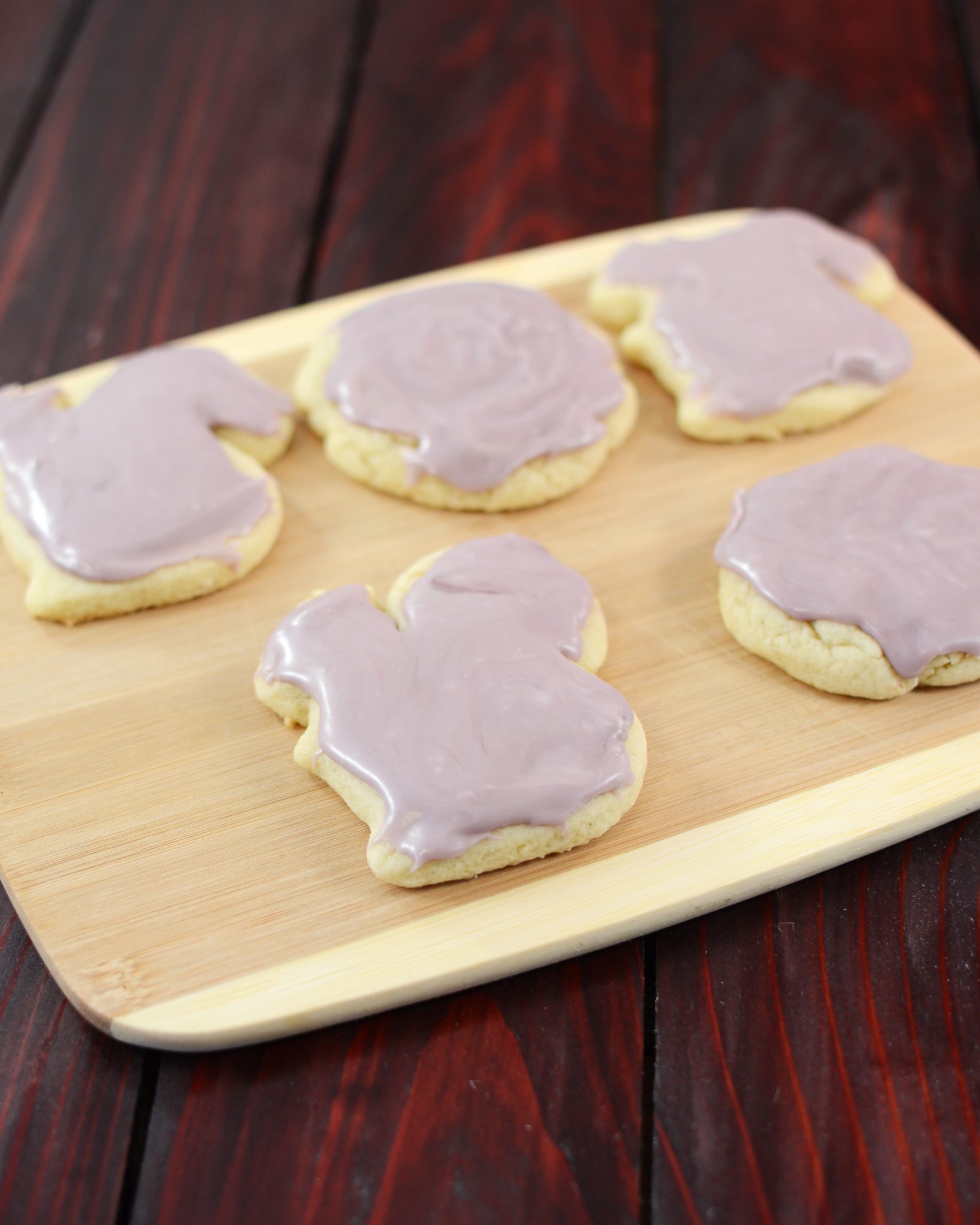 Five sugar cookies with purple frosting on a wooden cutting board.