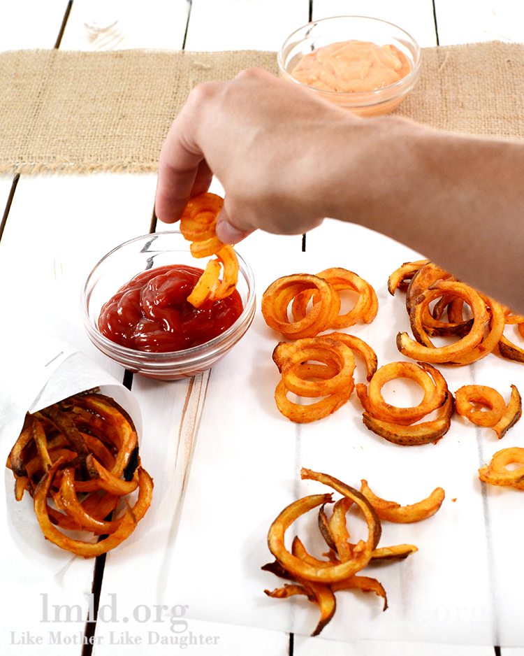 Angled view of oven baked curly fries on a wood board being dipped into ketchup.