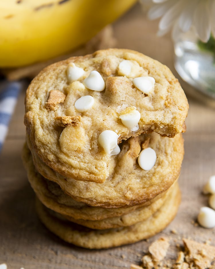 A stack of 4 banana cream pie cookies that are full of and topped with white chocolate chips and graham cracker pieces. There are bananas in the background of the photo. The top cookie has a bite taken out of it.