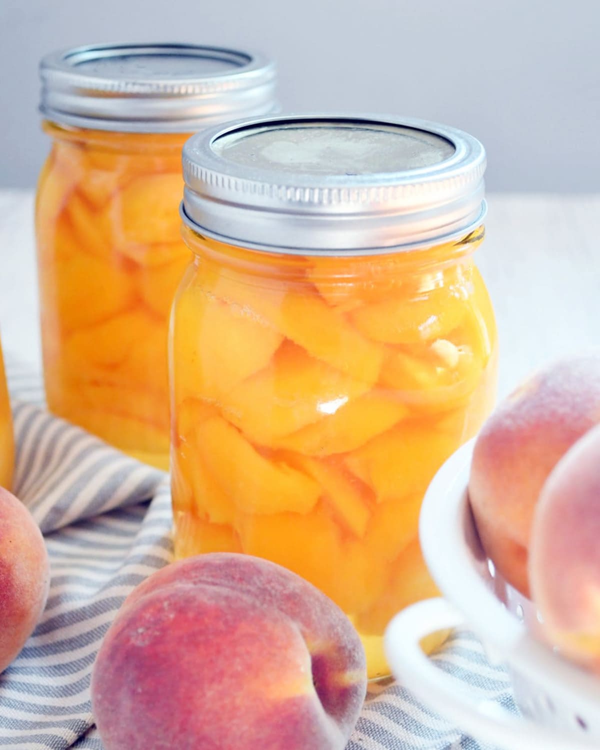 Peaches in a pint jar with lid, 2 pint jars with a few other whole peaches on a white table.