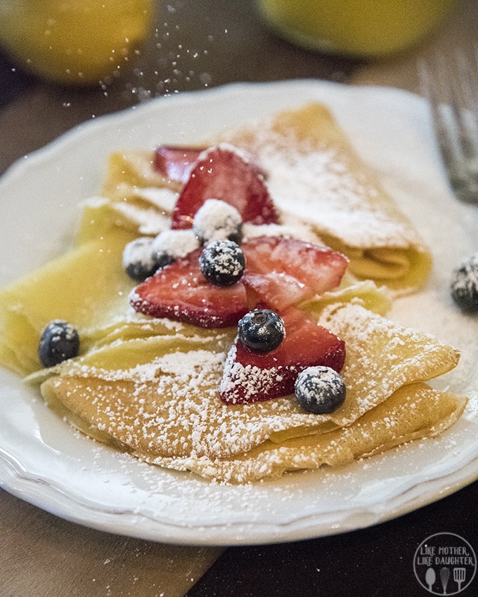 Crepes folded into squares and topped with powdered sugar, strawberries, and blueberries. 