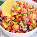 A close up of a bowl of peach salsa with red onion, tomatoes, jalapeno, and cilantro. The bowl has a chip in the salsa.