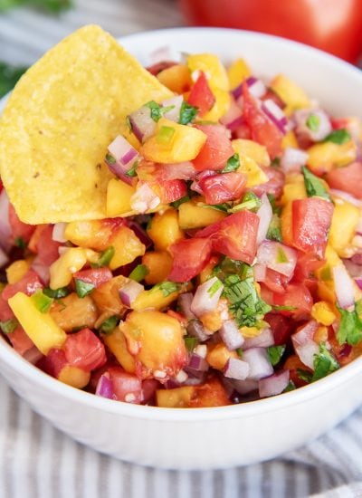 A close up of a bowl of peach salsa with red onion, tomatoes, jalapeno, and cilantro. The bowl has a chip in the salsa.
