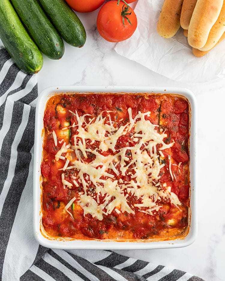 A white square baking dish full of zucchini lasagna shot from above. You can see the red sauce and sprinkles of baked shredded cheese over the top.