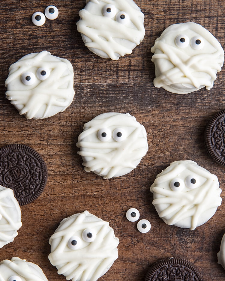 Mummy Oreos on a wooden board, they are white chocolate dipped Oreos decorated with candy eyes and drizzled to look like mummies.