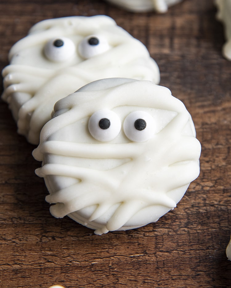 Mummy Oreos on a wooden board, they are white chocolate dipped Oreos decorated with candy eyes and drizzled to look like mummies.