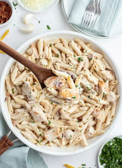 A Pasta dish in a pan with penne noodles and chicken in a white sauce. Topped with a sprinkle of parsley.