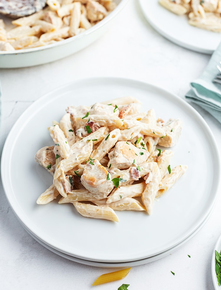 An alfredo pasta on a white plate, with penne noodles, chicken pieces, and a white sauce, the top is sprinkled with chopped parsley.