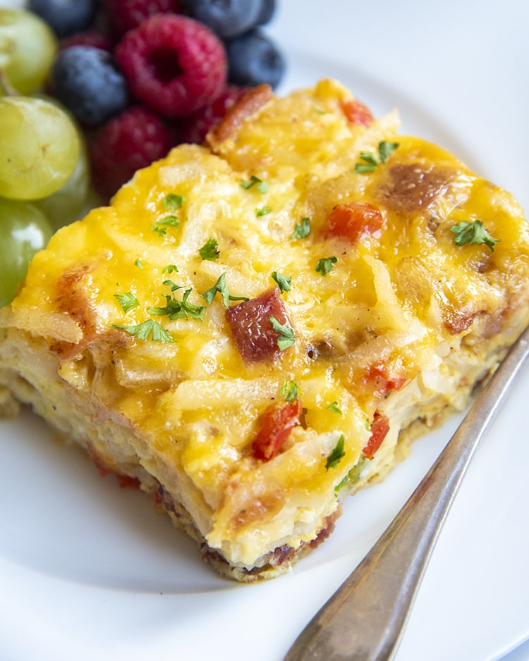 A piece of a savory breakfast casserole on a white plate, with grapes and fresh berries behind it.