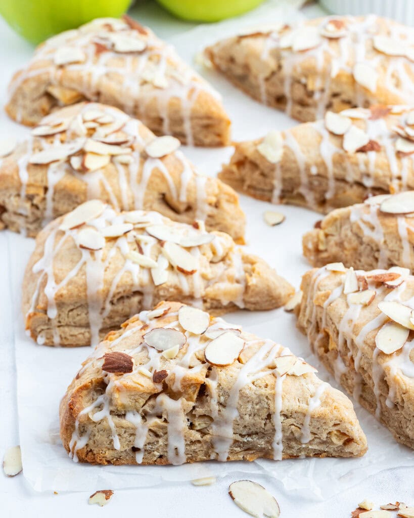 A white counter top topped with almond apple scones, with a glaze, and sliced almonds on top.