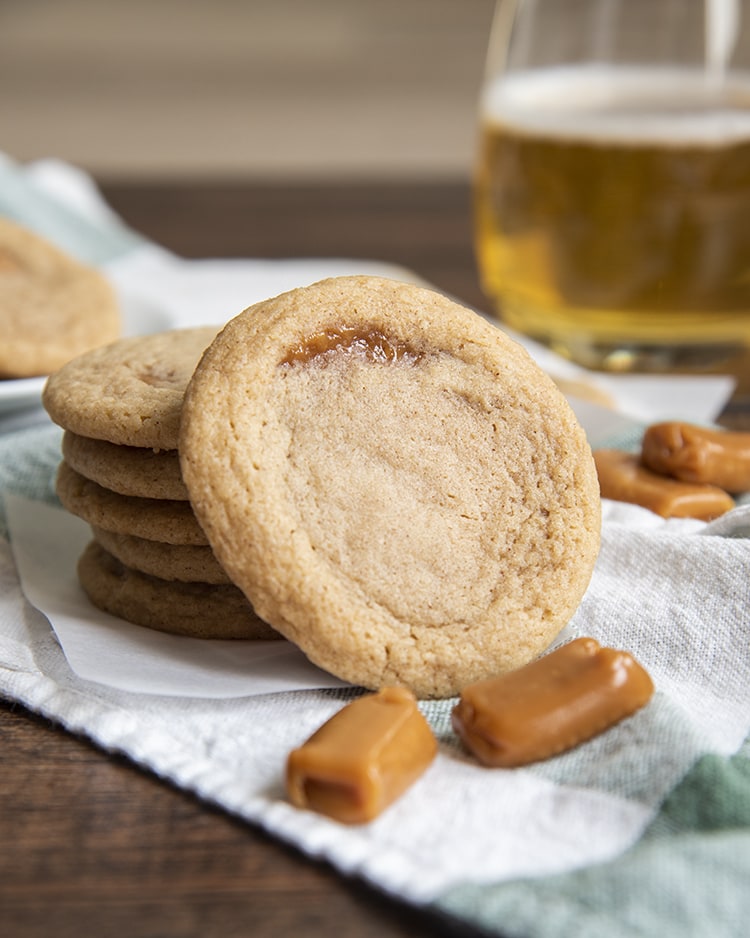 A caramel apple cider cookie leaning against a stack of cookies with two caramels on the ground in front of it.