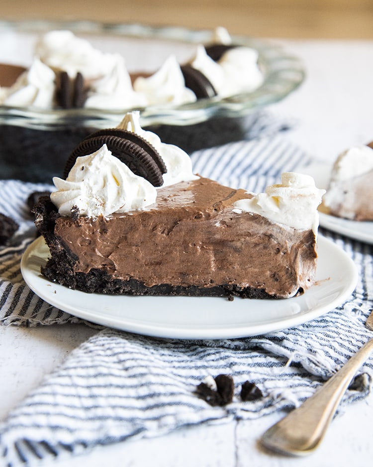 A side shot of a slice of chocolate cream pie. It has an Oreo crust and is topped with whipped cream rosettes.