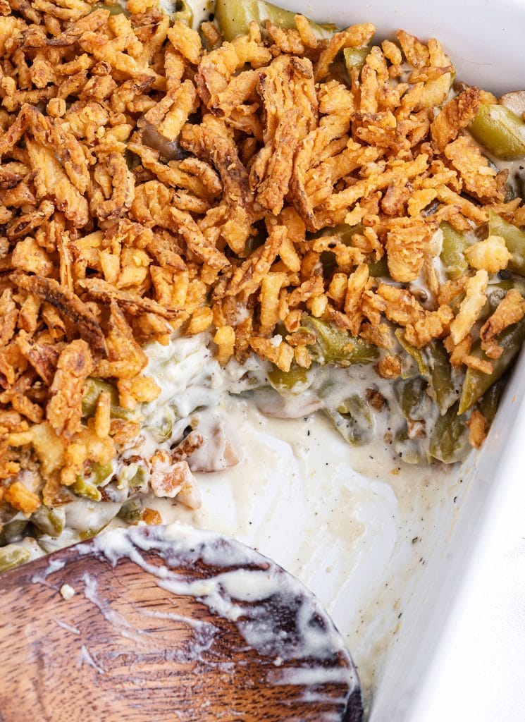 A pan of green bean casserole topped with friend onion straws with a wooden spoon in the pan.