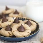 A small blue plate topped with a few peanut butter blossoms leaning on each other, with a cup of milk behind the plate.