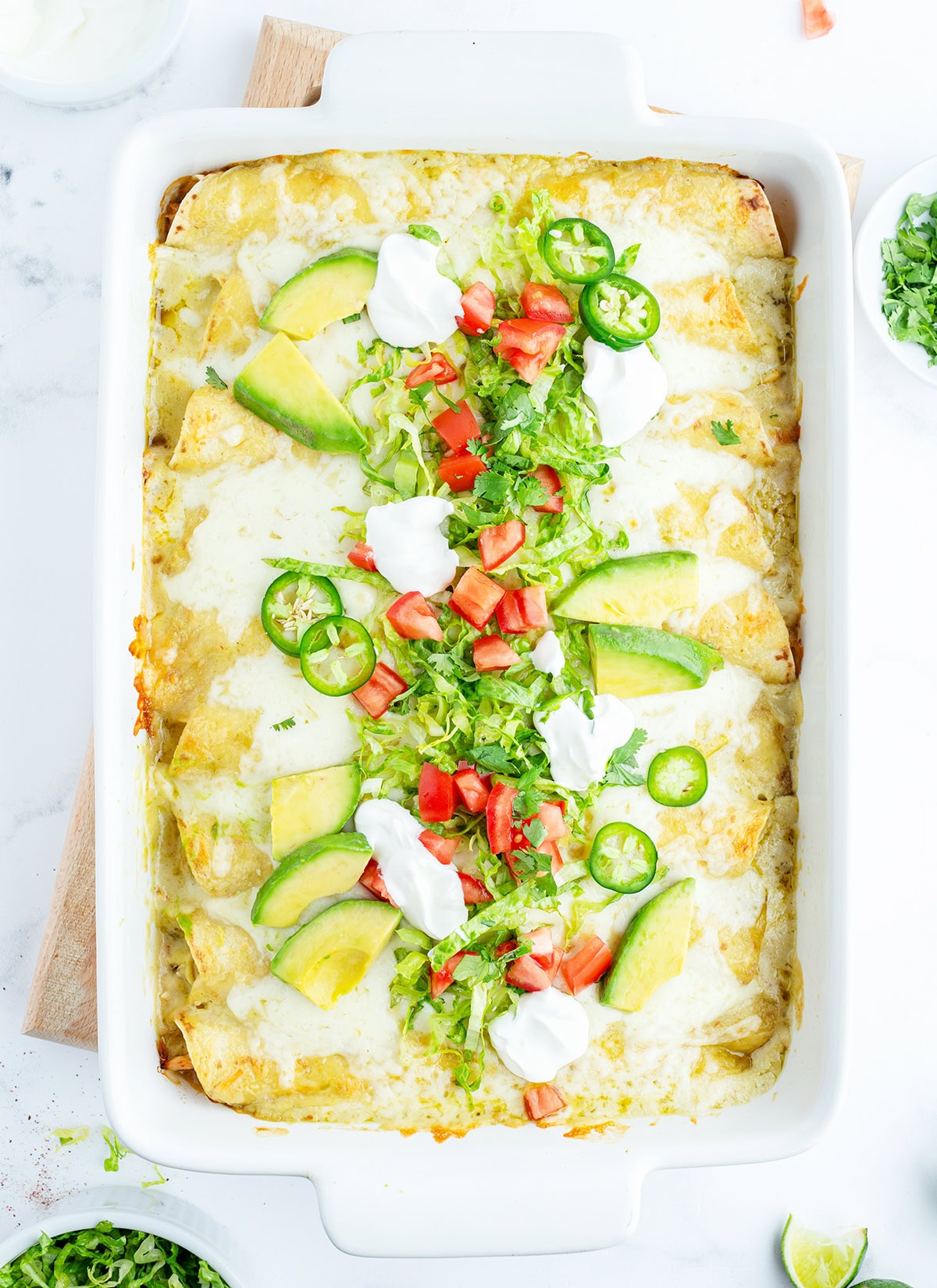 A pan of chicken enchiladas topped with avocado slices, tomatoes, and jalapenos.