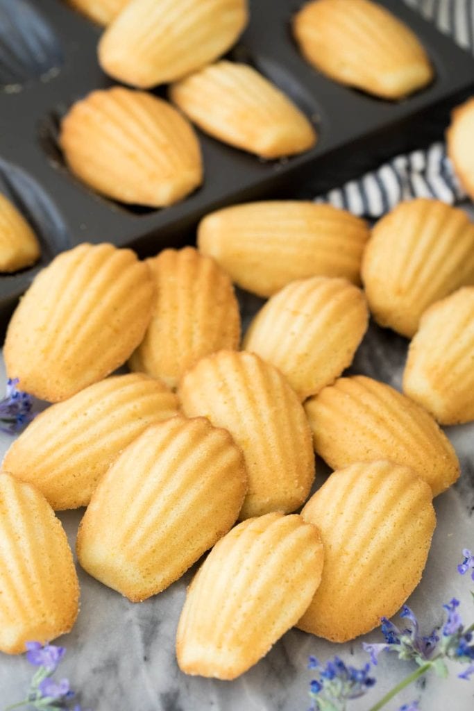 A pile of madeleine cookies in front of a pan filled with more of them.