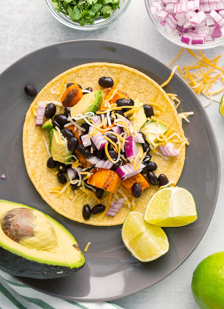 A sweet potato and black bean taco on a plate with an avocado and lime on the side.