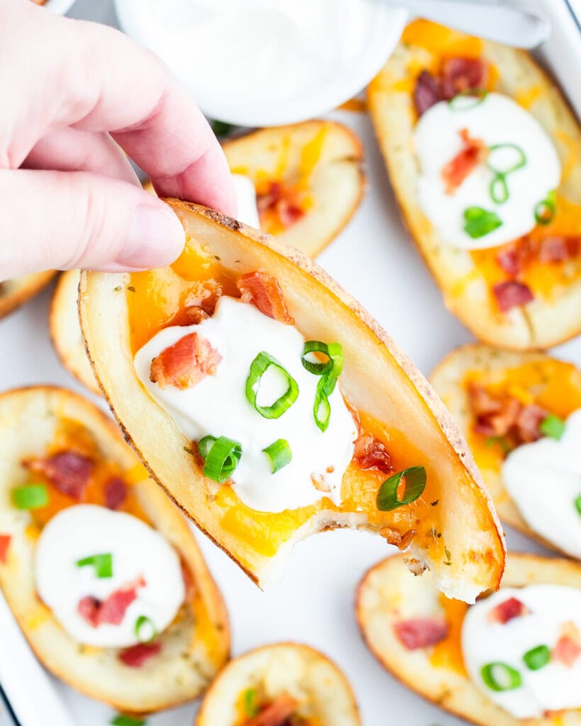 A hand holding a crispy potato skin topped with cheese, sour cream, and bacon, with a bite out of it.