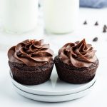 Two chocolate cupcakes with chocolate frosting on small white plates. There are bottles of milk behind them.