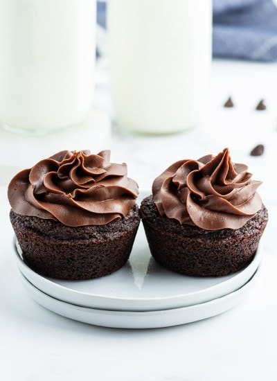 Two chocolate cupcakes with chocolate frosting on small white plates. There are bottles of milk behind them.