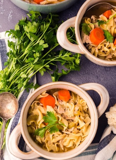 Above image of chicken noodle soup in white bowls with ingredients surrounding them.