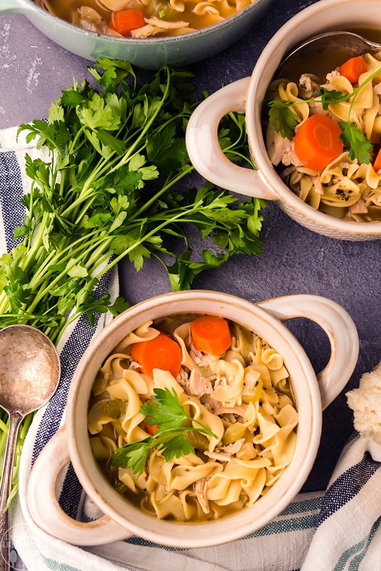 Above image of chicken noodle soup in white bowls with ingredients surrounding them.