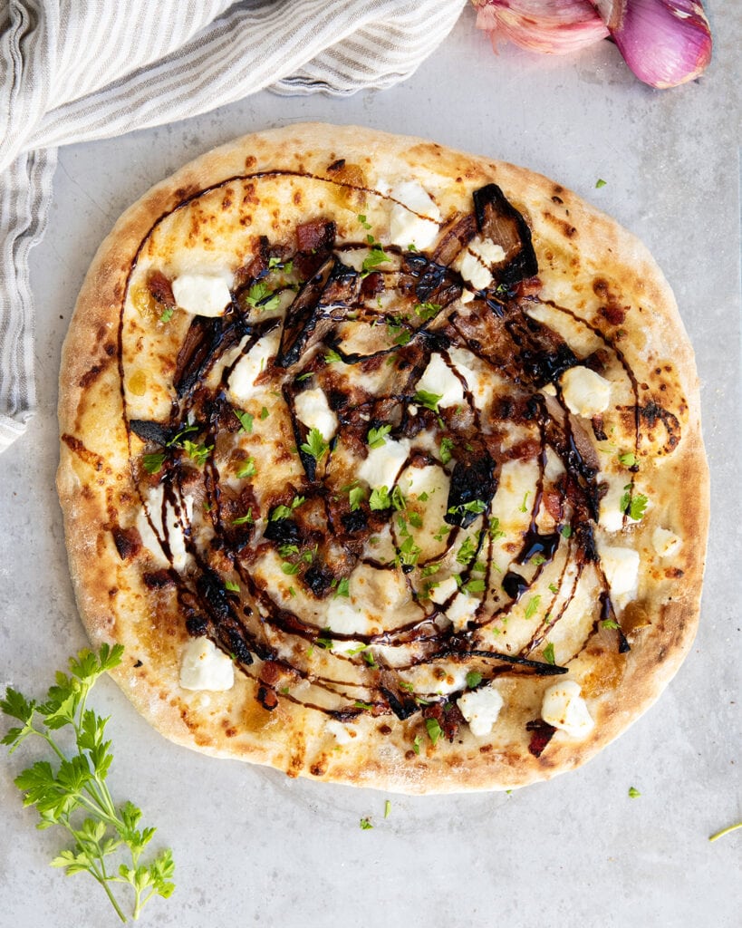 An above view of a pizza topped with goat cheese, sauteed scallions, bacon, and balsamic glaze.