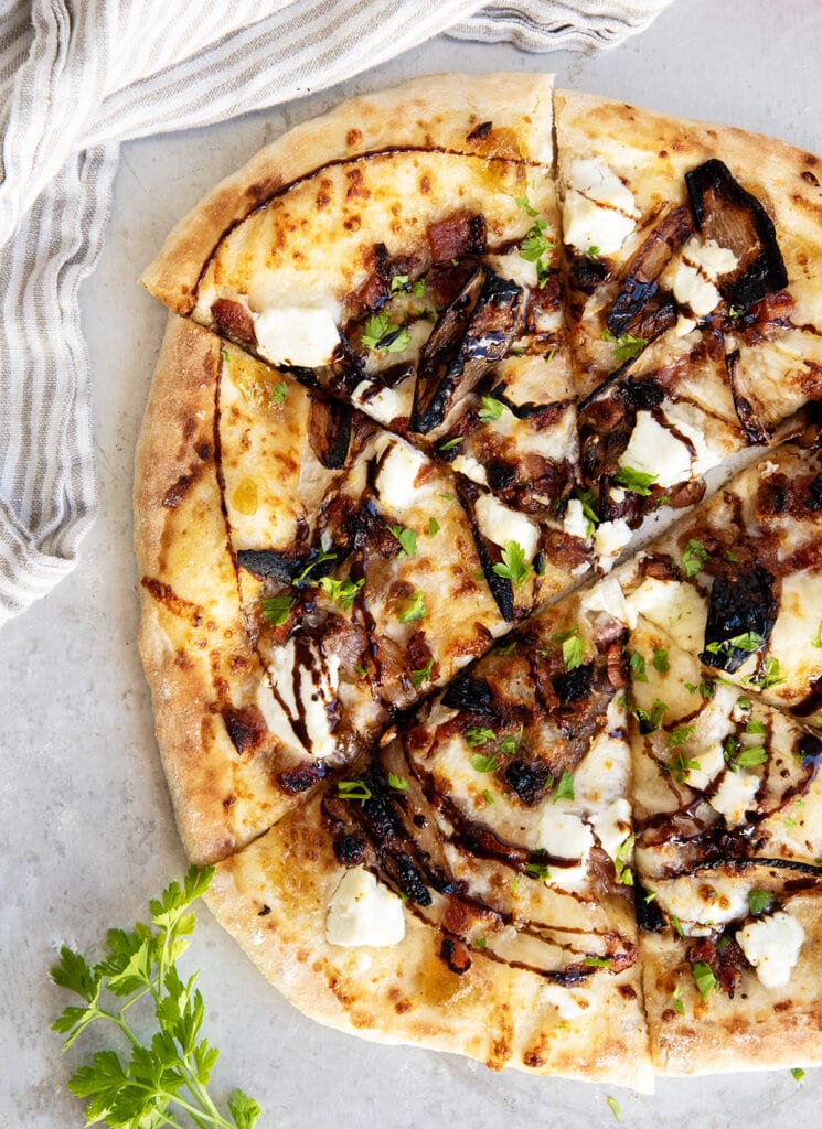An above view of a sliced pizza topped with goat cheese, sauteed scallions, bacon, and balsamic glaze.