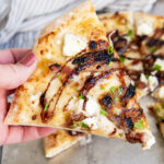 A hand holding a piece of goat cheese pizza topped with balsamic glaze.