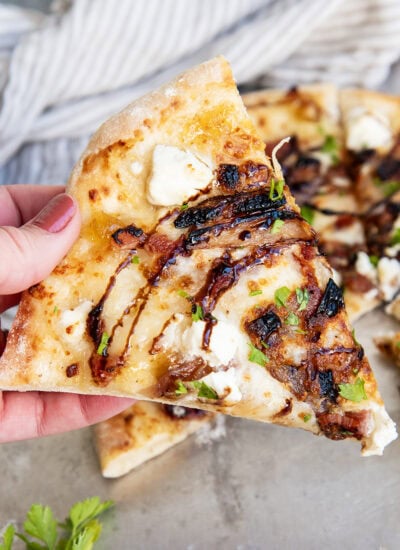 A hand holding a piece of goat cheese pizza topped with balsamic glaze.