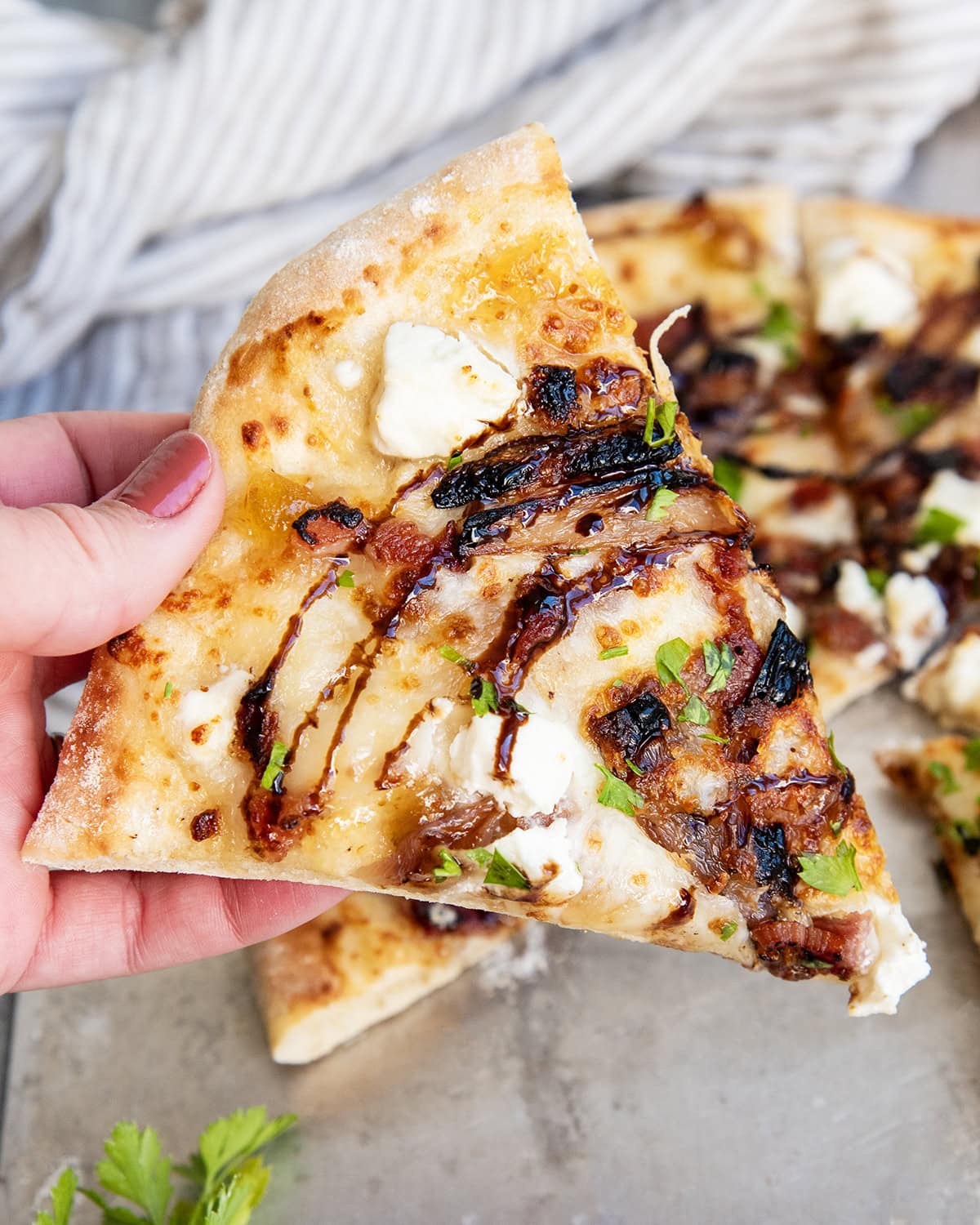 A hand holding a piece of goat cheese pizza topped with balsamic glaze.