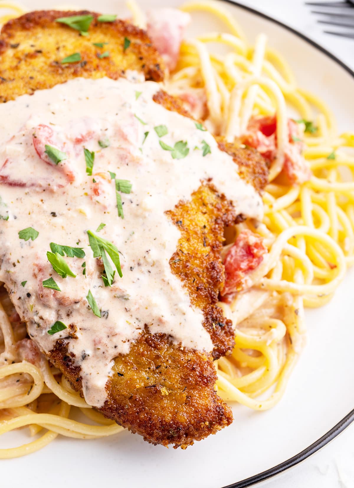 A close up of a breaded chicken breast on spaghetti noodles. It's topped with a pink tomato sauce and fresh cut parsley.