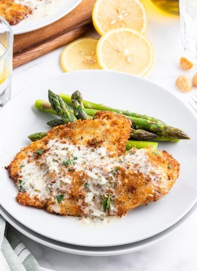 Tilapia on a plate with a golden brown crust covering it, and it is topped with a creamy white herb sauce, and laying next to asparagus on a plate.