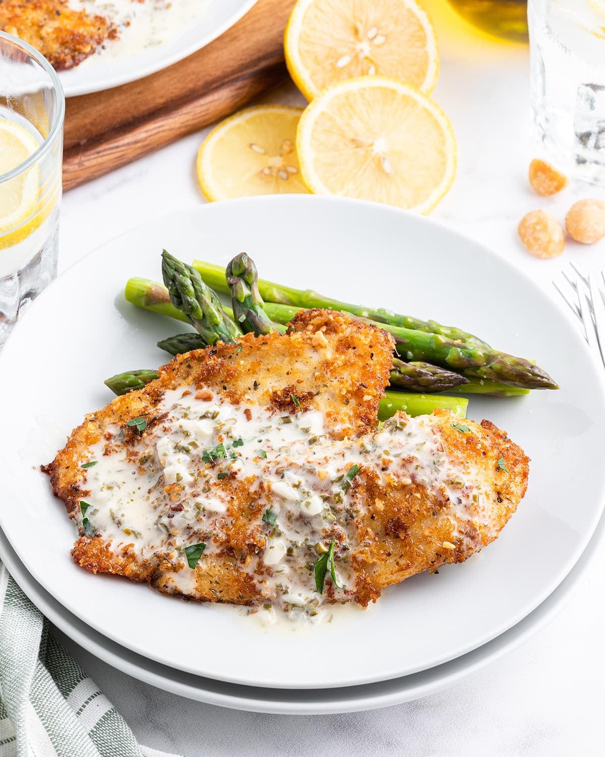Tilapia on a plate with a golden brown crust covering it, and it is topped with a creamy white herb sauce, and laying next to asparagus on a plate.