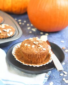 pumpkin oat muffin in an open paper liner on a brown plate along side a butter knife with butter, orange pumpkin and muffin tin in background