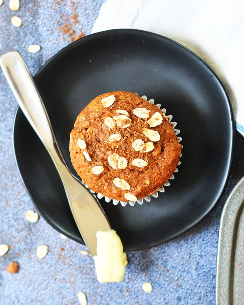pumpkin oat muffin on a brown plate with a butter knife and butter beside it, oats sprinkled around