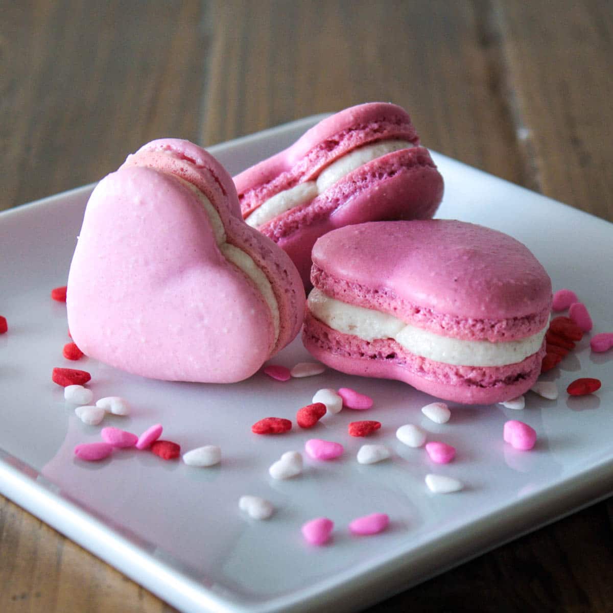 A close up shot of three valentine\'s day heart macarons on a plate with heart shaped sprinkles.
