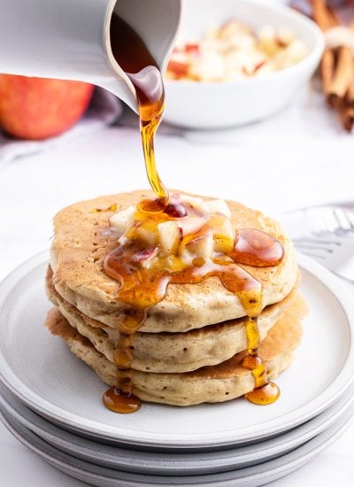 A stack of pancakes topped with diced apples and syrup is being poured over the top.