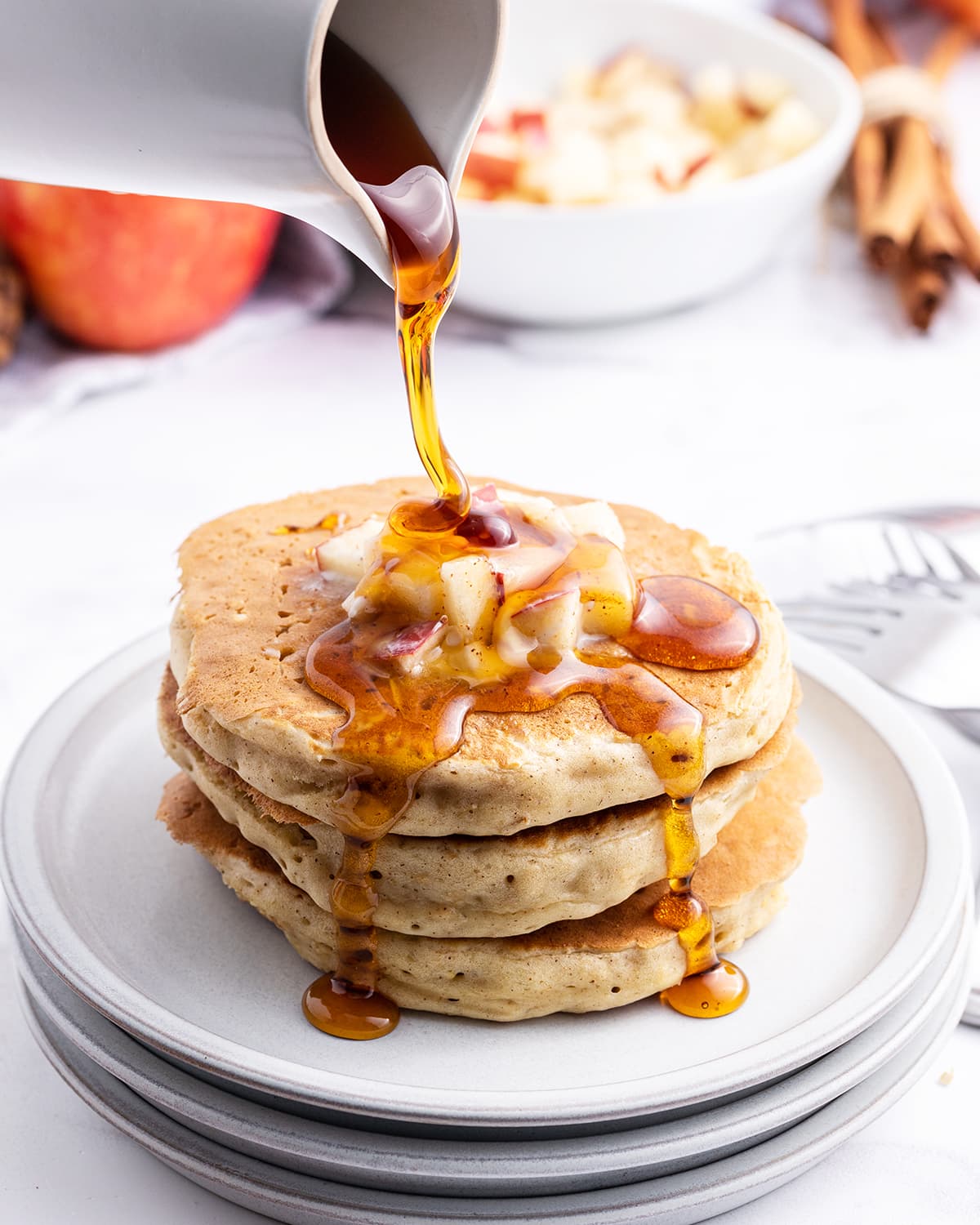 A stack of pancakes topped with diced apples and syrup is being poured over the top.