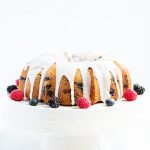 Lemon Berry Bundt Cake with a lemon glaze on a white cake stand with fresh berries on the side