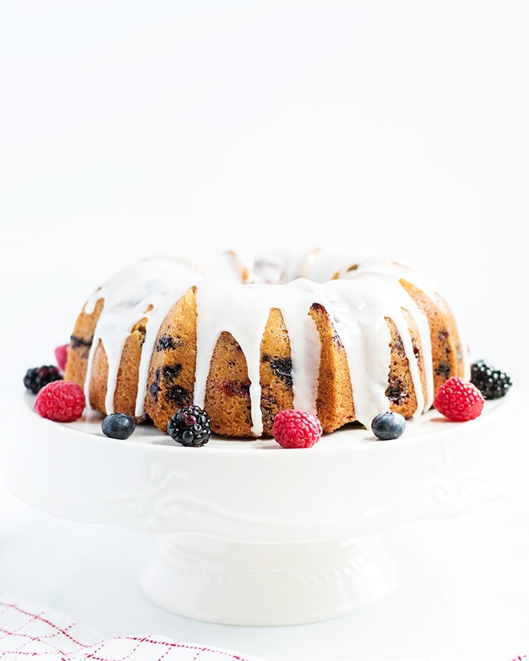Lemon Berry Bundt Cake with a lemon glaze on a white cake stand with fresh berries on the side