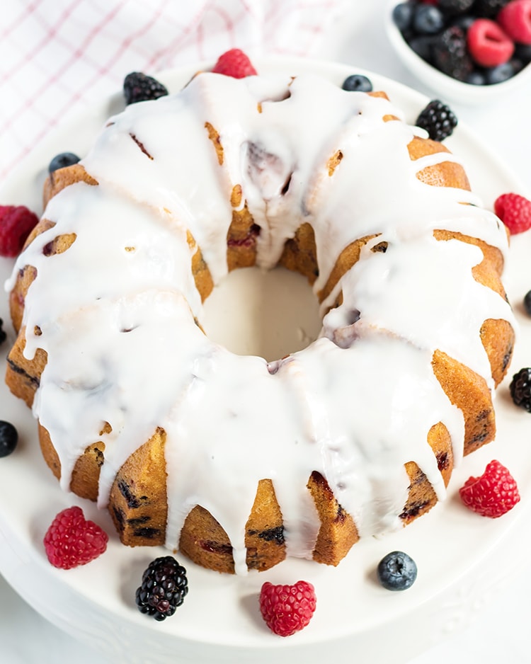 Lemon Berry Bundt Cake with a lemon glaze on a white cake stand with fresh berries on the side
